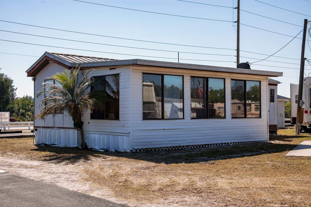 White mobile home with large windows, palm tree, and telephone poles in a sunny, outdoor setting.