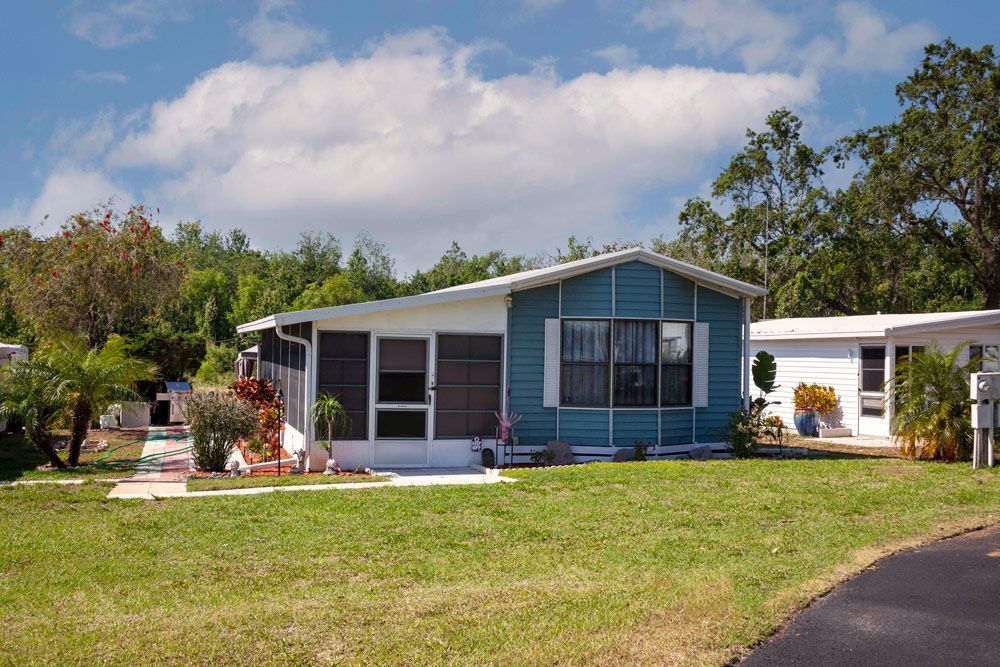 Blue and white mobile home with a screened-in porch and green lawn under a sunny sky.