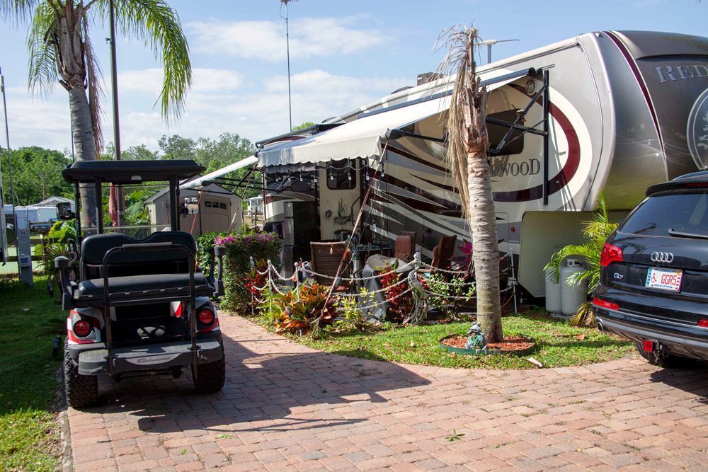 RV parked on brick, with golf cart and SUV. Awning extended, surrounded by plants and outdoor seating.