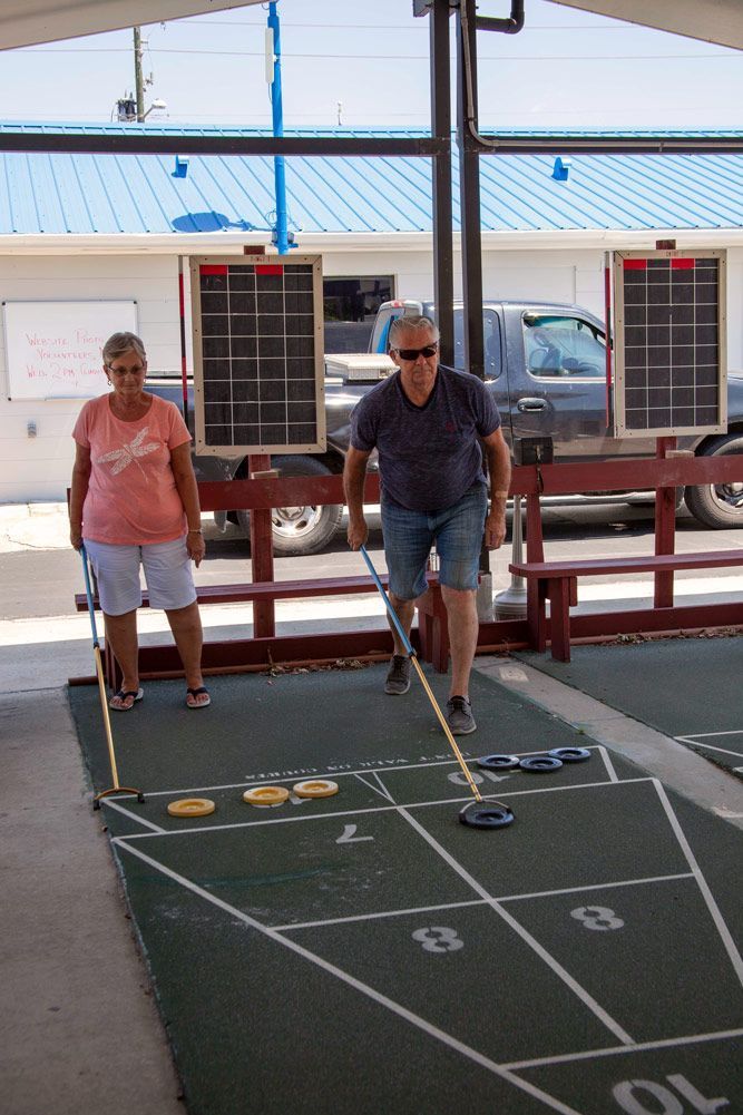 Two people playing shuffleboard on a court; man in shorts pushes a disc, woman watches.