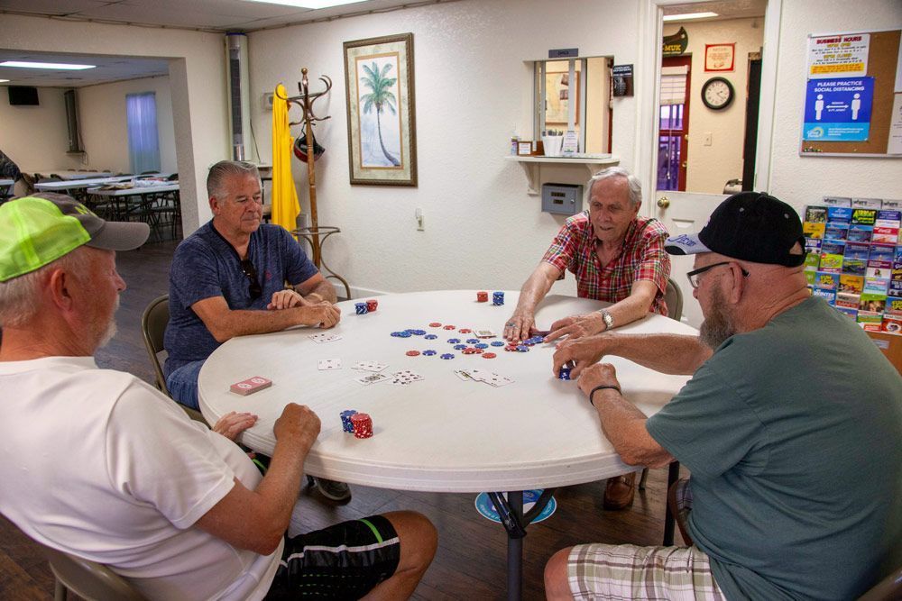 Four men seated around a white table playing a game with chips and cards in a community center.