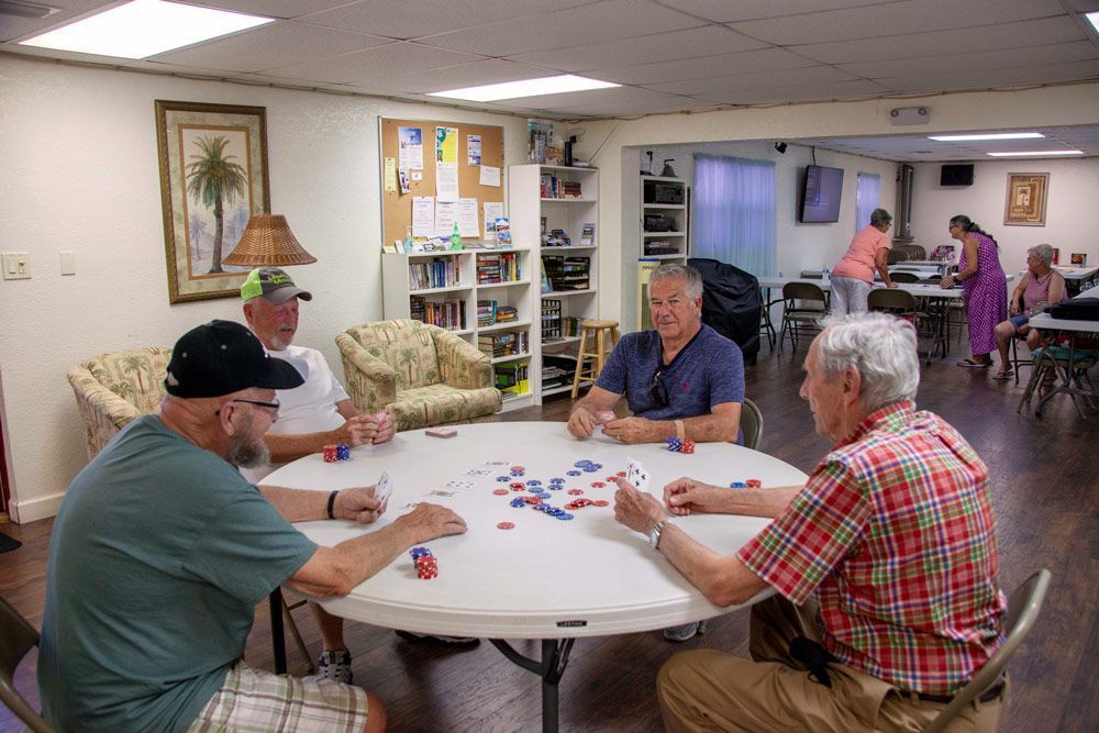 Four men playing cards at a round table indoors. Two other people stand nearby.