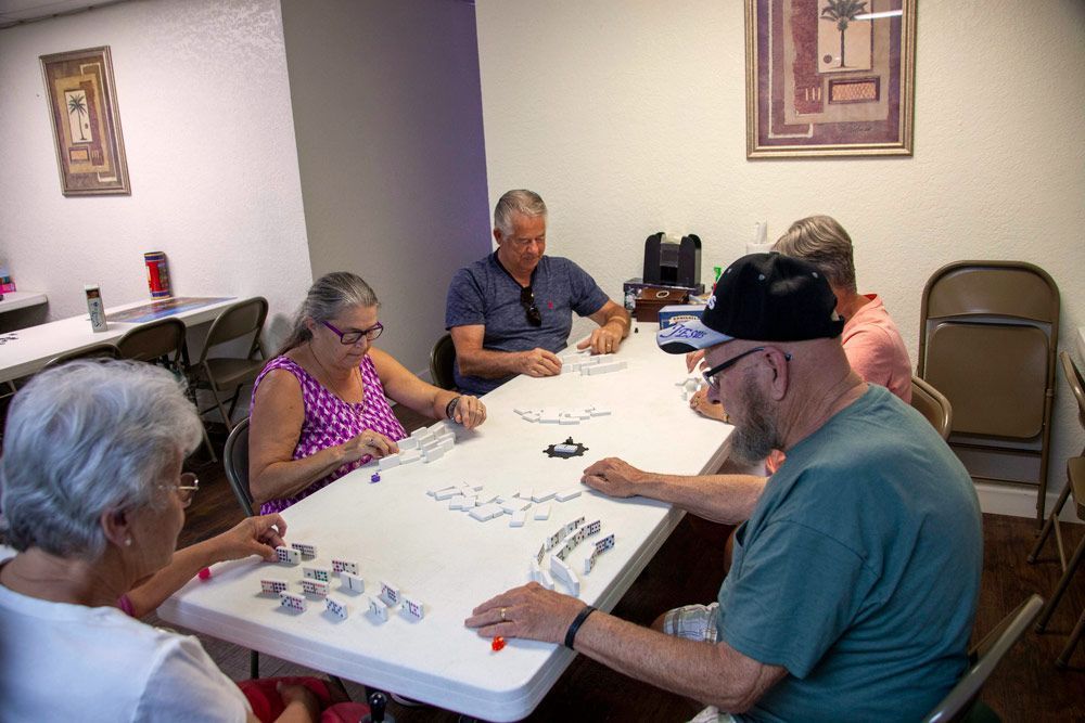 Five people playing dominoes at a table in a room with neutral-colored walls, chairs, and two framed pictures.