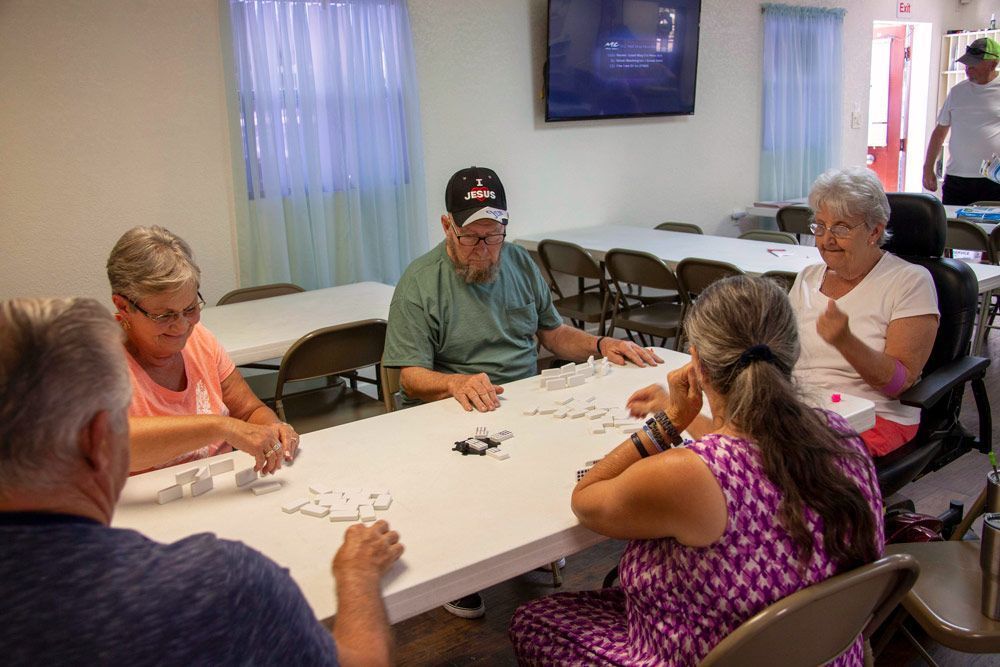 People playing dominoes at a table in a room. Some smile. A TV hangs on the wall.