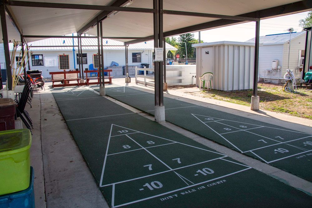 Shuffleboard courts under a covered outdoor area. Green courts with white markings. Light gray concrete and posts.