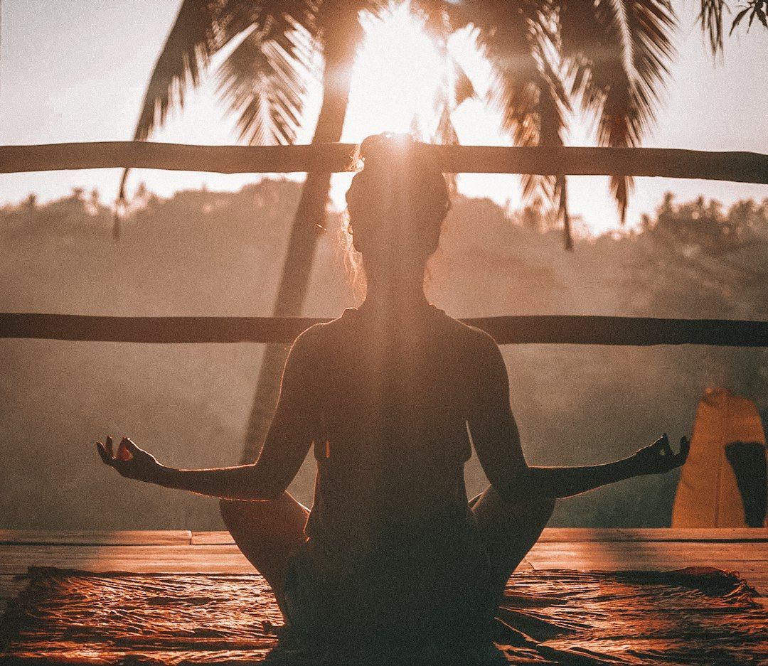 A woman is sitting in a lotus position on a wooden deck at sunset.