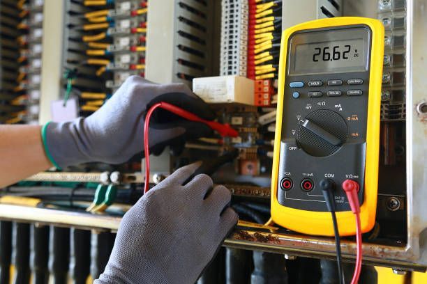 An electrician wearing gloves tests a control panel with a multimeter, displaying 26.62.