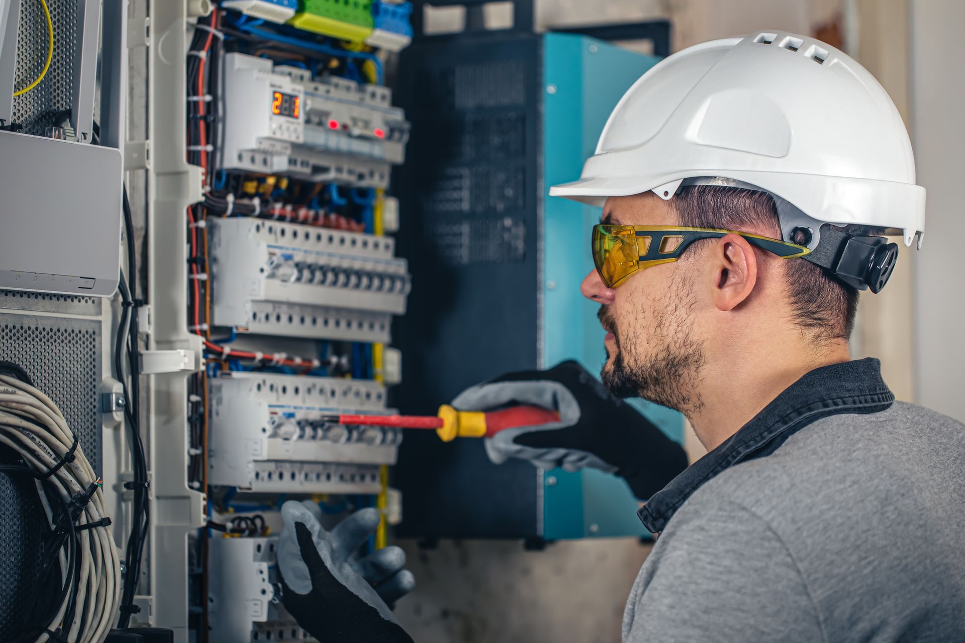 Electrician in a hard hat and safety glasses working on a fuse box.  He is using a screwdriver, wearing gloves, and inspecting electrical wiring.