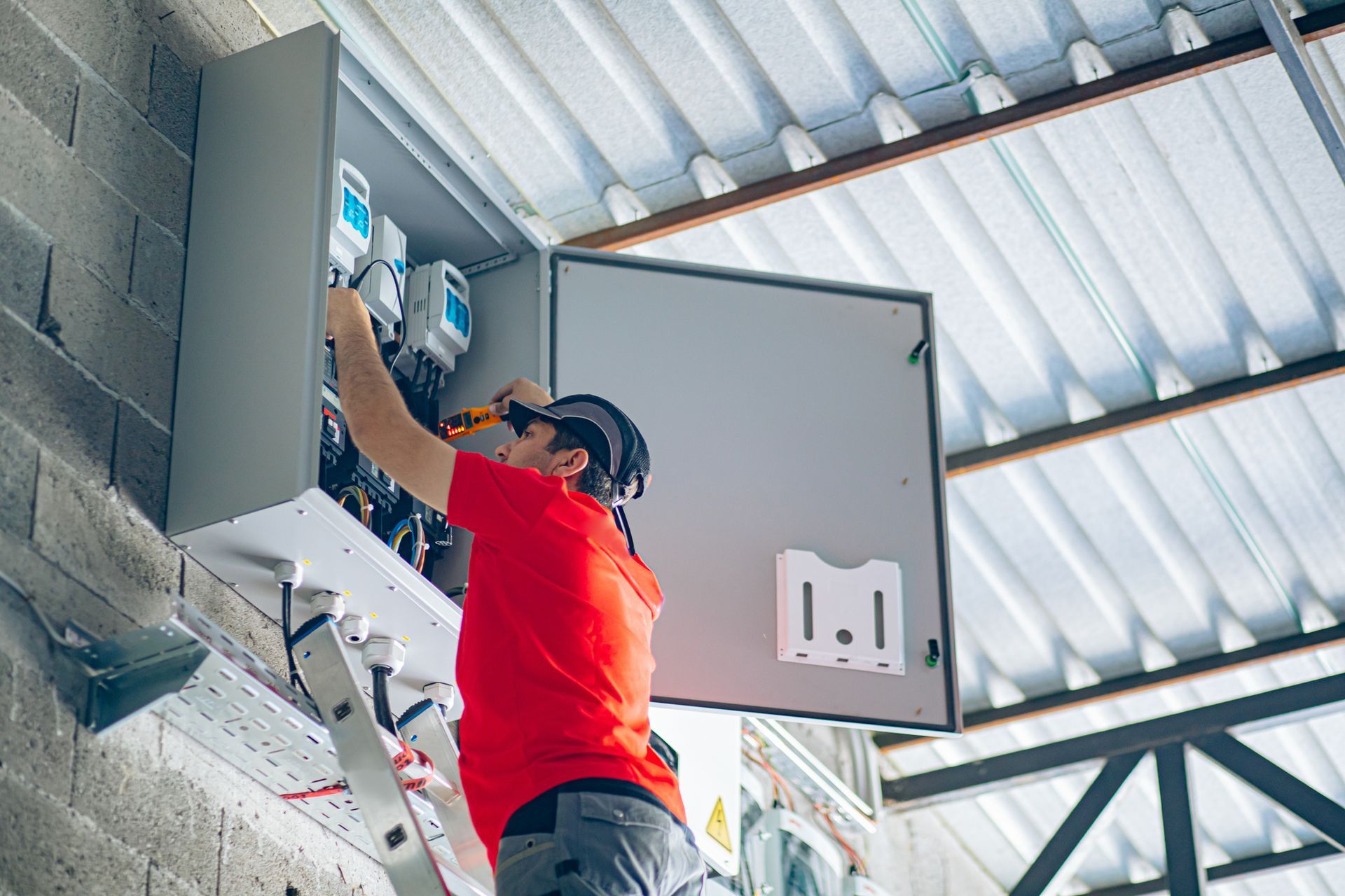 An electrician in a red shirt works on an electrical panel mounted on a cinder block wall, using a screwdriver.