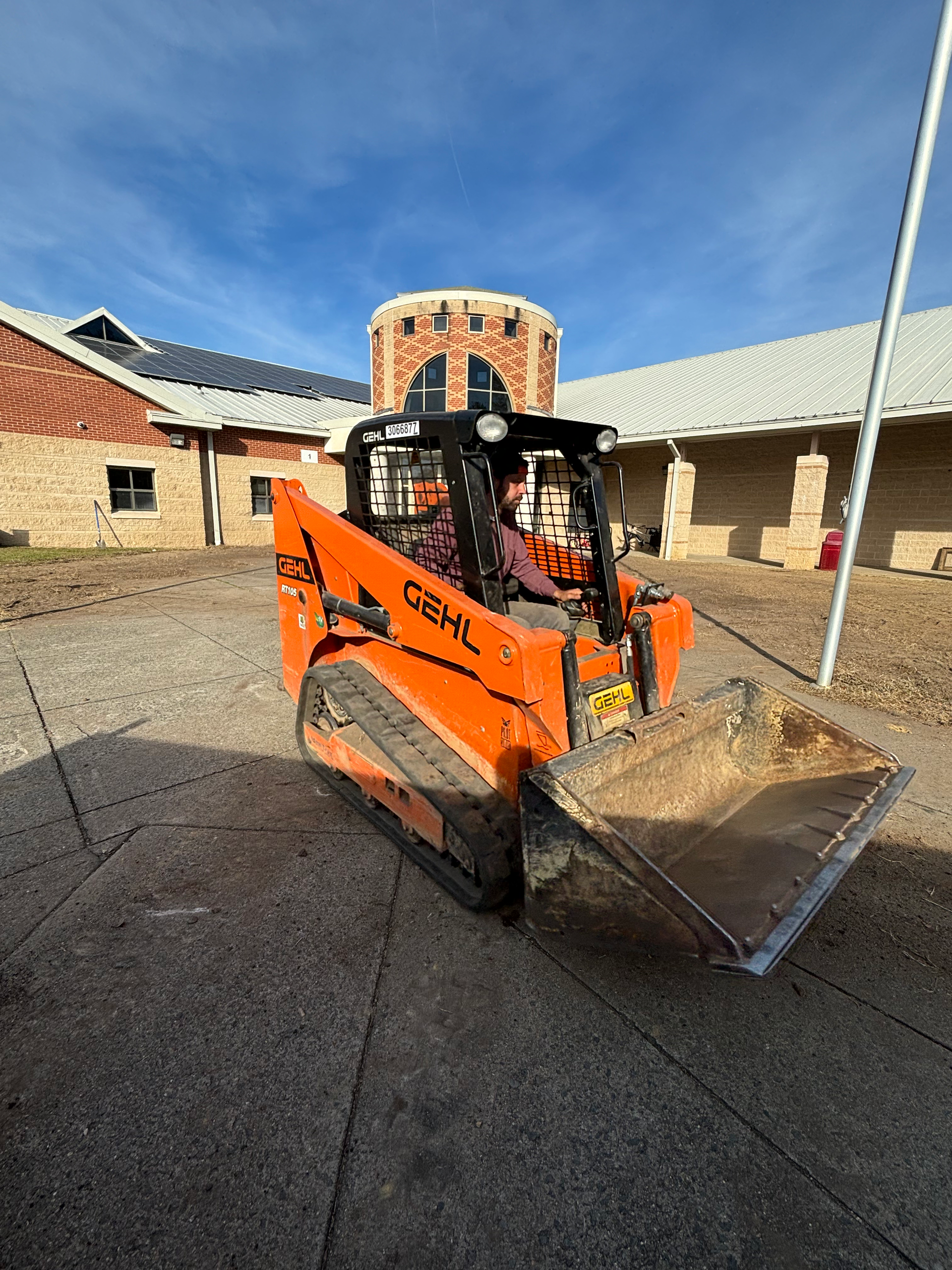Orange Gehl skid steer loader with full bucket, near a brick building.