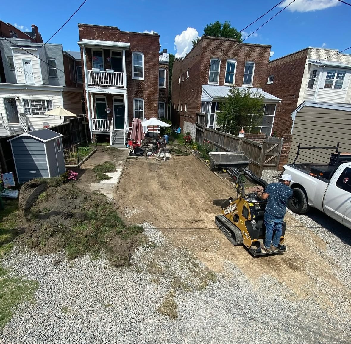 A person operating a compact track loader on a dirt lot between two brick houses.
