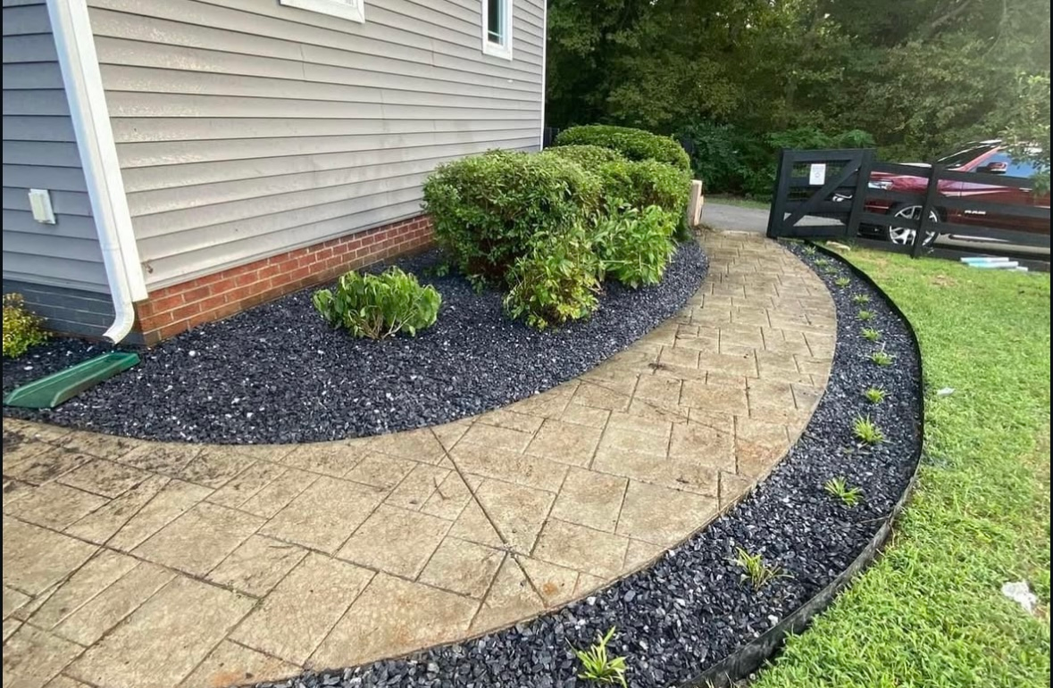 Curved walkway with stamped concrete and black rock border, alongside house and landscaping.