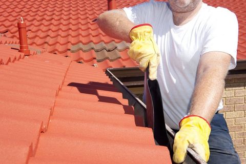 Handyman, worker cleaning gutter on house with shovel, roof with red tiles and shingles as background.