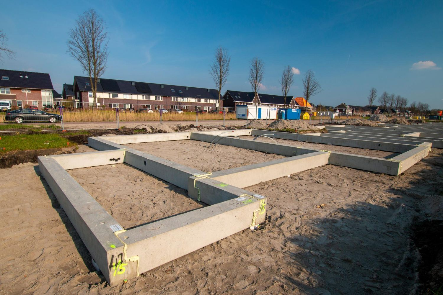 Concrete foundations laid out on a sandy construction site with houses in the background.