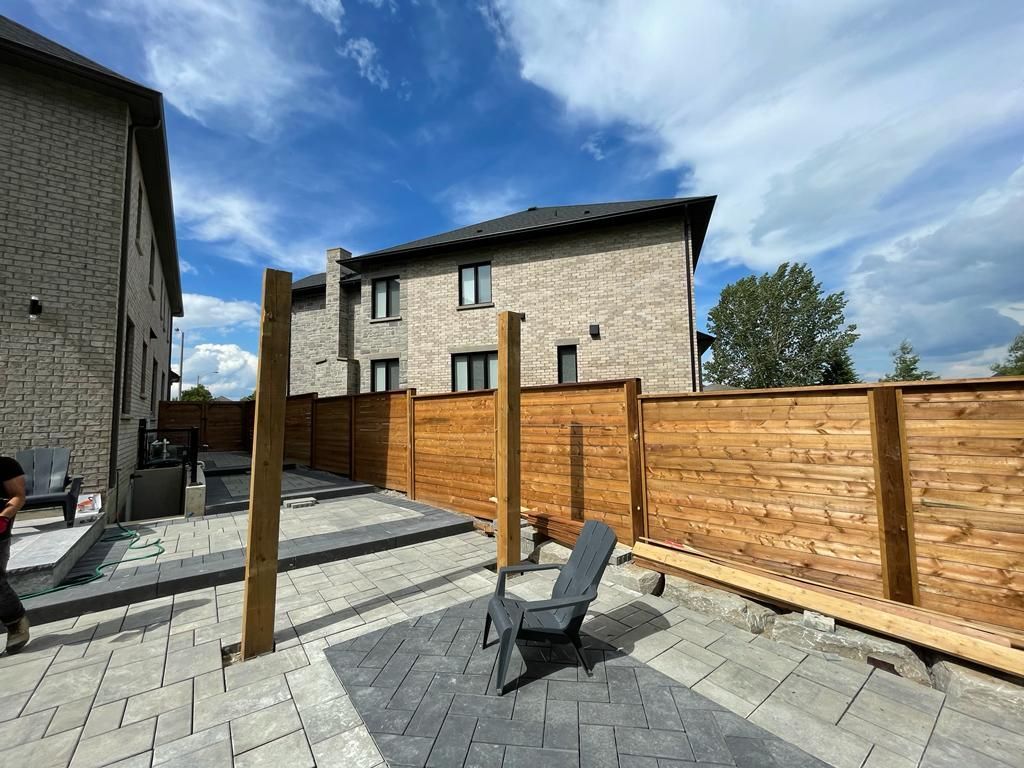 Backyard with wooden fence, brick buildings, gray patio stones, blue sky with clouds. Chair in the center.