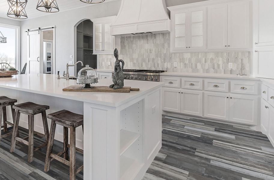 White kitchen with island and wooden stools, gray and white tiled backsplash, and gray flooring.