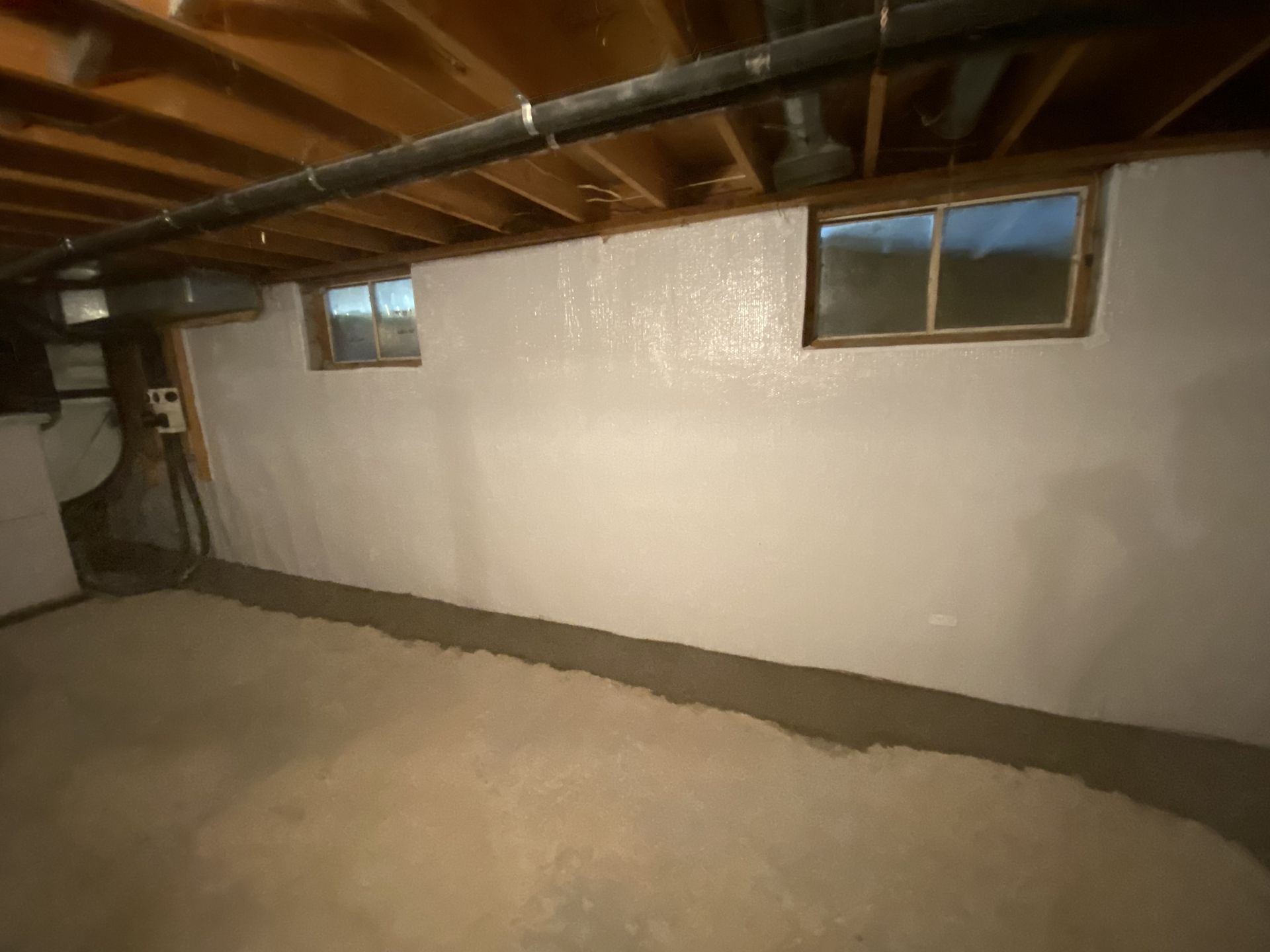 Basement interior with white painted walls, two small windows, exposed ceiling beams, and concrete floor.