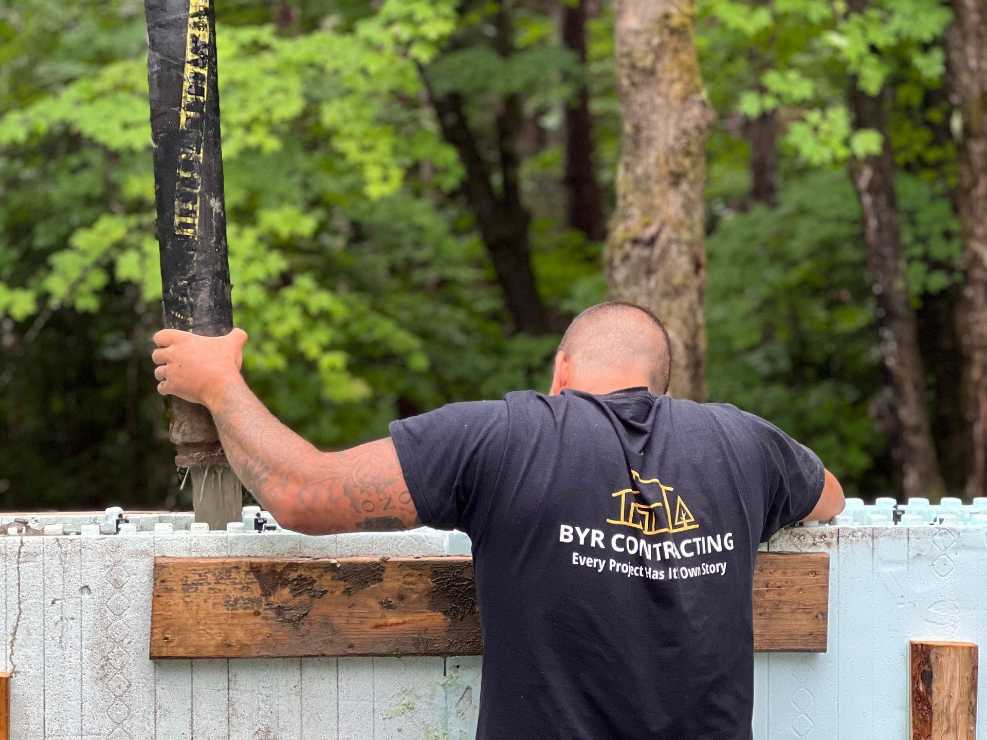 A man wearing a black shirt that says byr contracting is standing in front of a wooden fence.