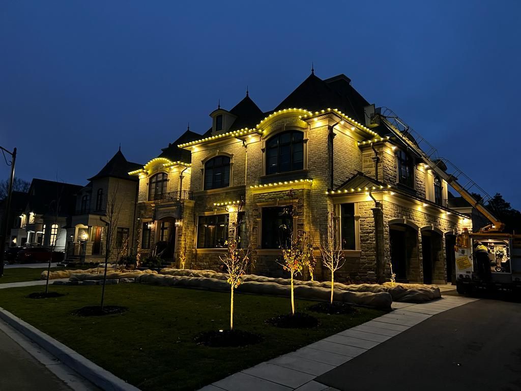 A large stone house at dusk, lit with yellow Christmas lights along the roofline and windows.