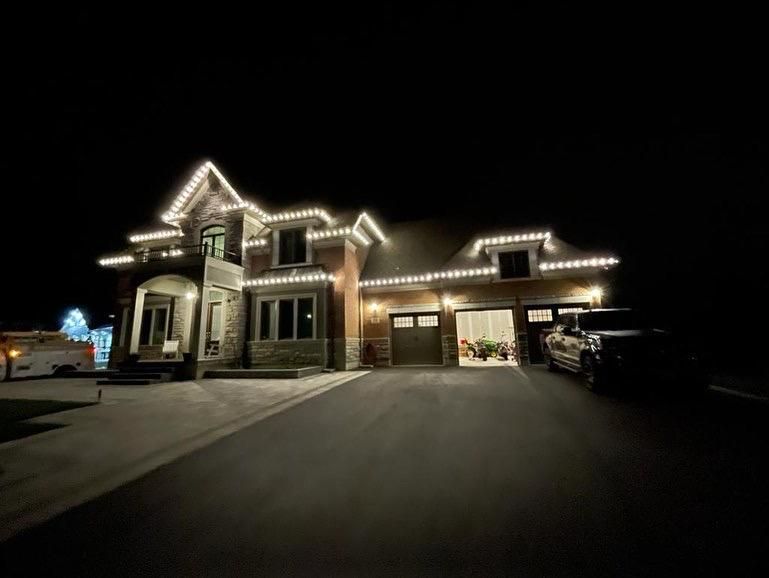 House exterior at night, lit with white Christmas lights outlining the roof and windows. A vehicle is parked in the driveway.