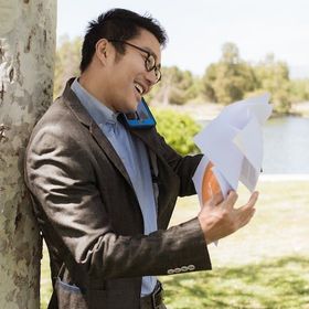 An image man leaning on a tree answering a phone while holding a bunch of papers