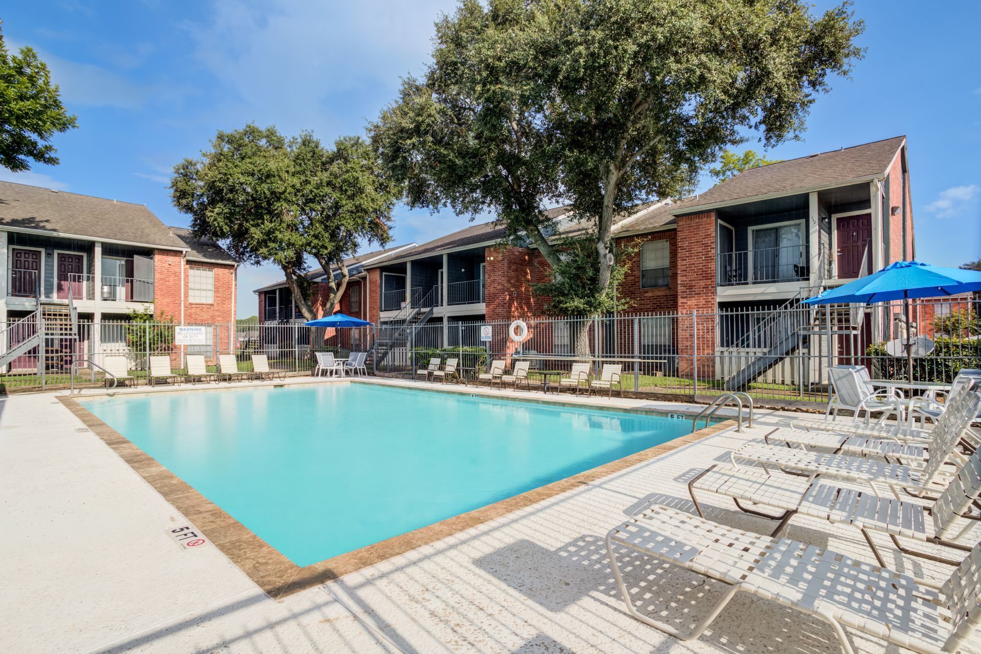 Apartment complex with a blue swimming pool, lounge chairs, and trees on a sunny day.