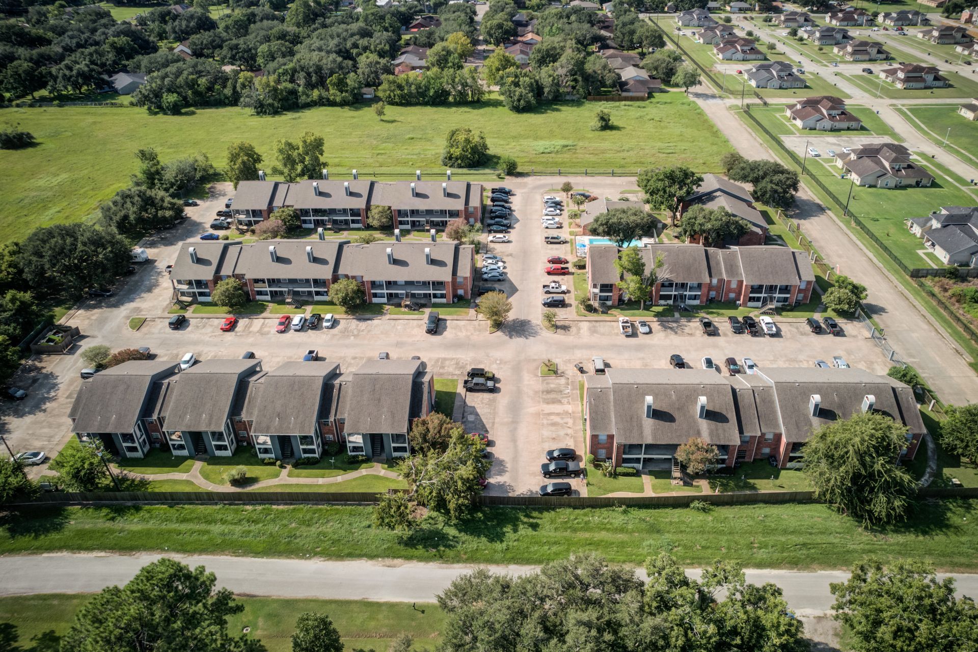 Aerial view of apartment complex with parking, a pool, and surrounding green space.