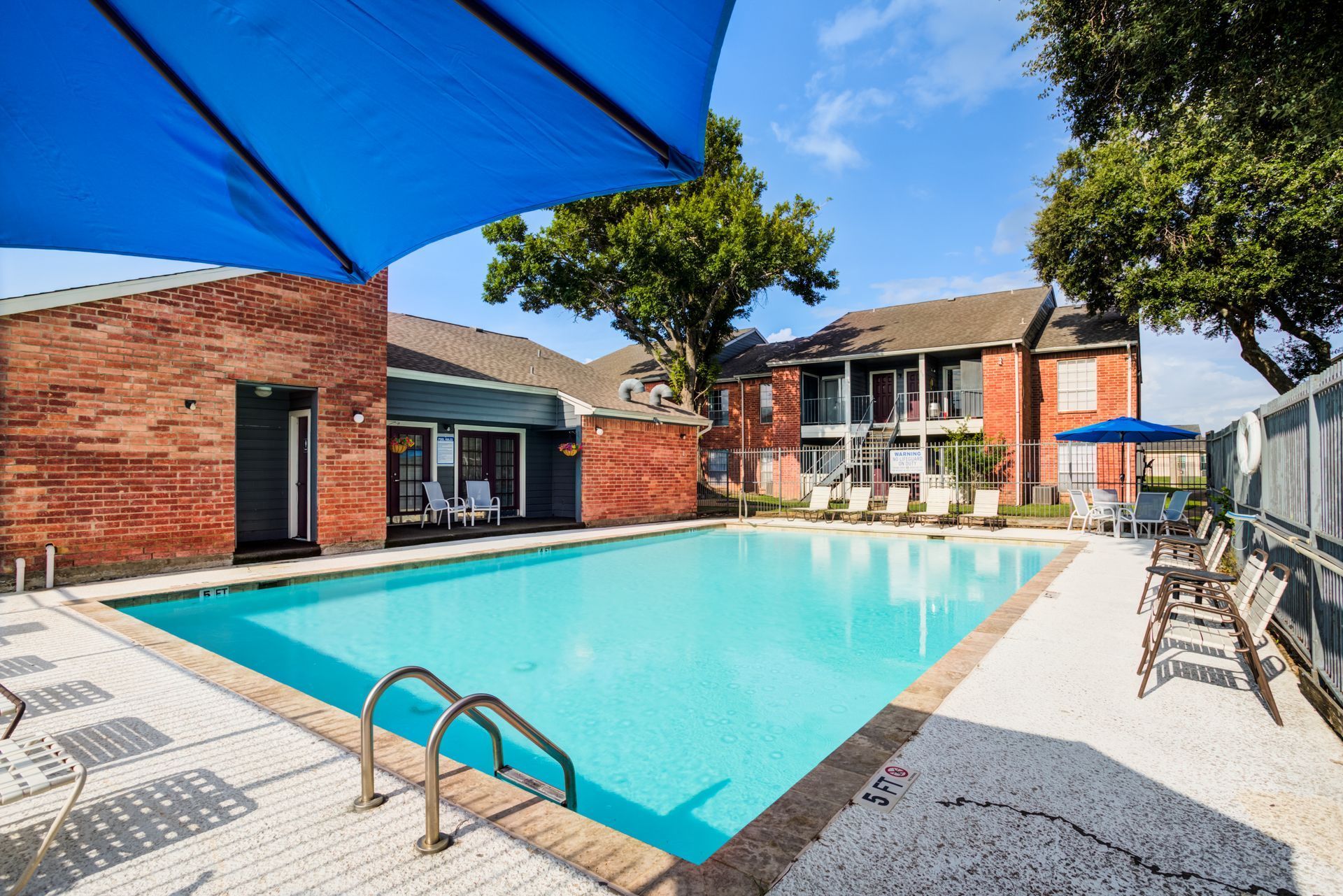 Pool with blue umbrella, surrounded by brick building and apartment complex on a sunny day.