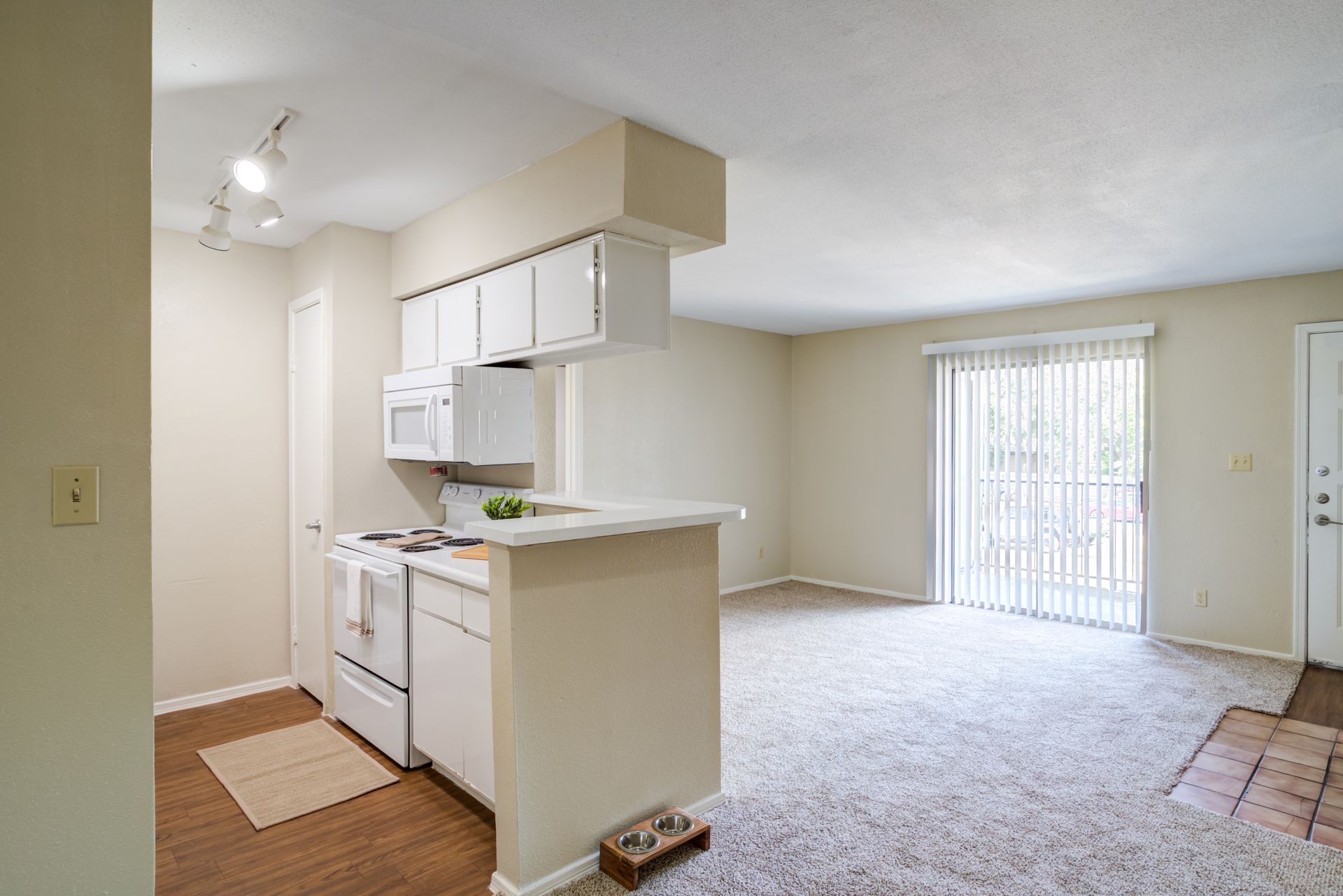 A kitchen with white appliances opens to a living room with sliding glass doors; carpet flooring.