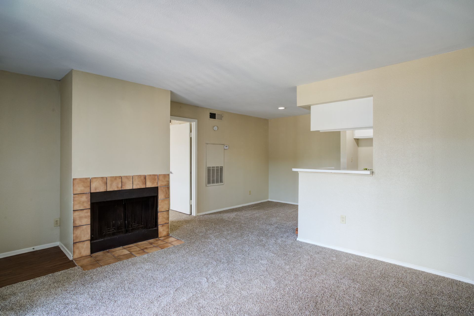 Empty living room with fireplace, kitchen pass-through, and beige walls and carpet.