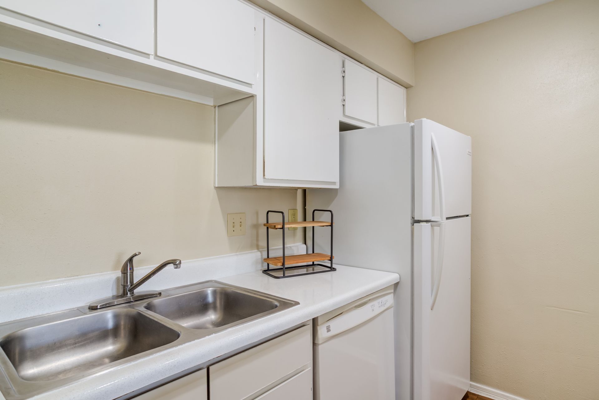 White kitchen with sink, cabinets, refrigerator, and small shelving unit.