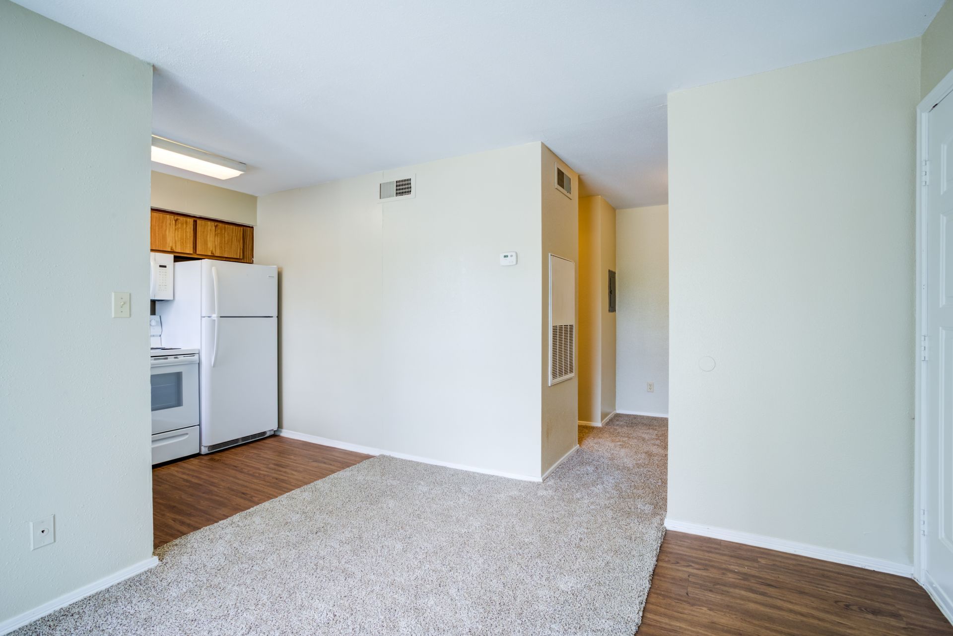 A small apartment's interior, showing a kitchen with white appliances and a living area with beige carpet.
