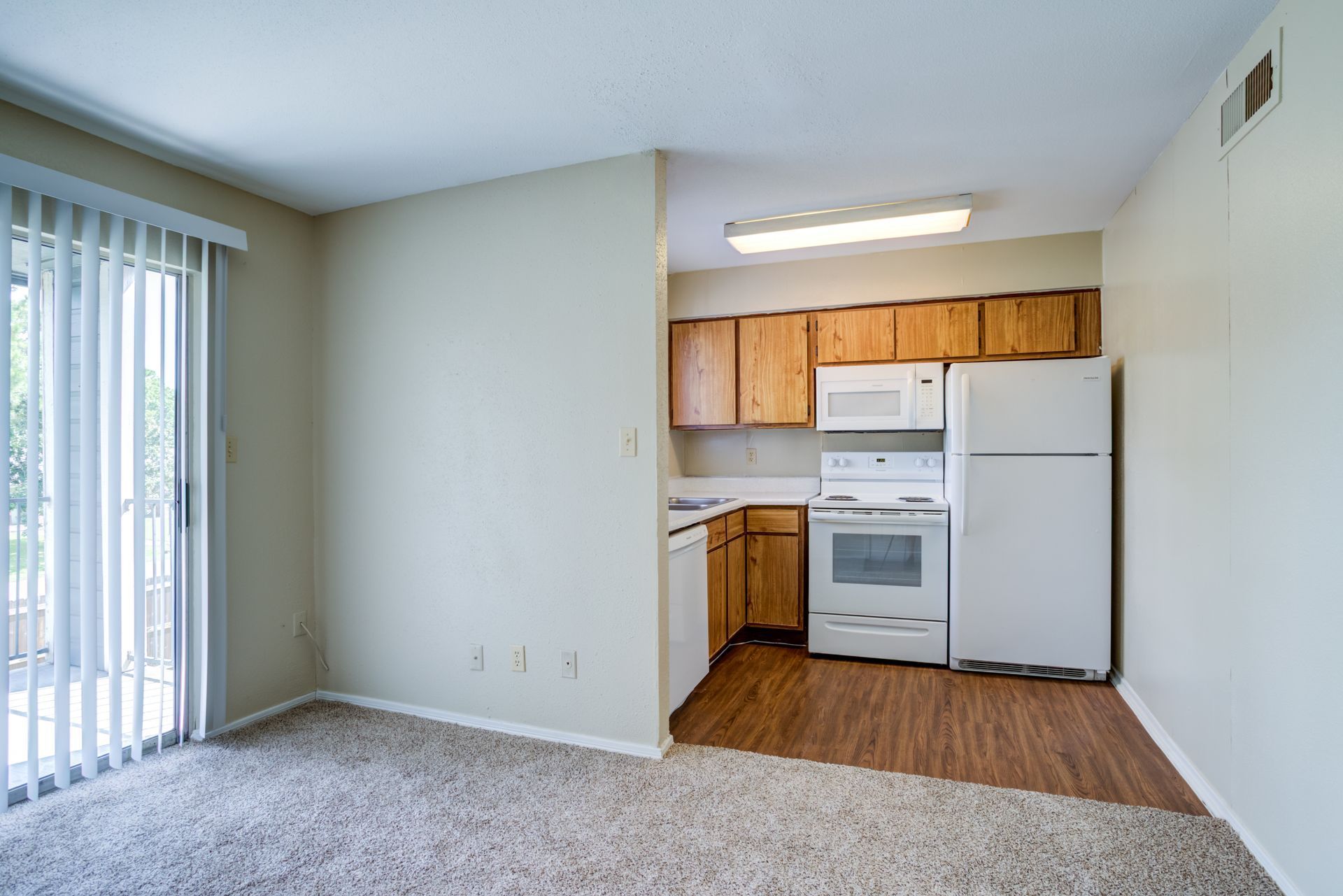Interior view of a kitchen and living room with brown cabinets, white appliances, and a sliding glass door.