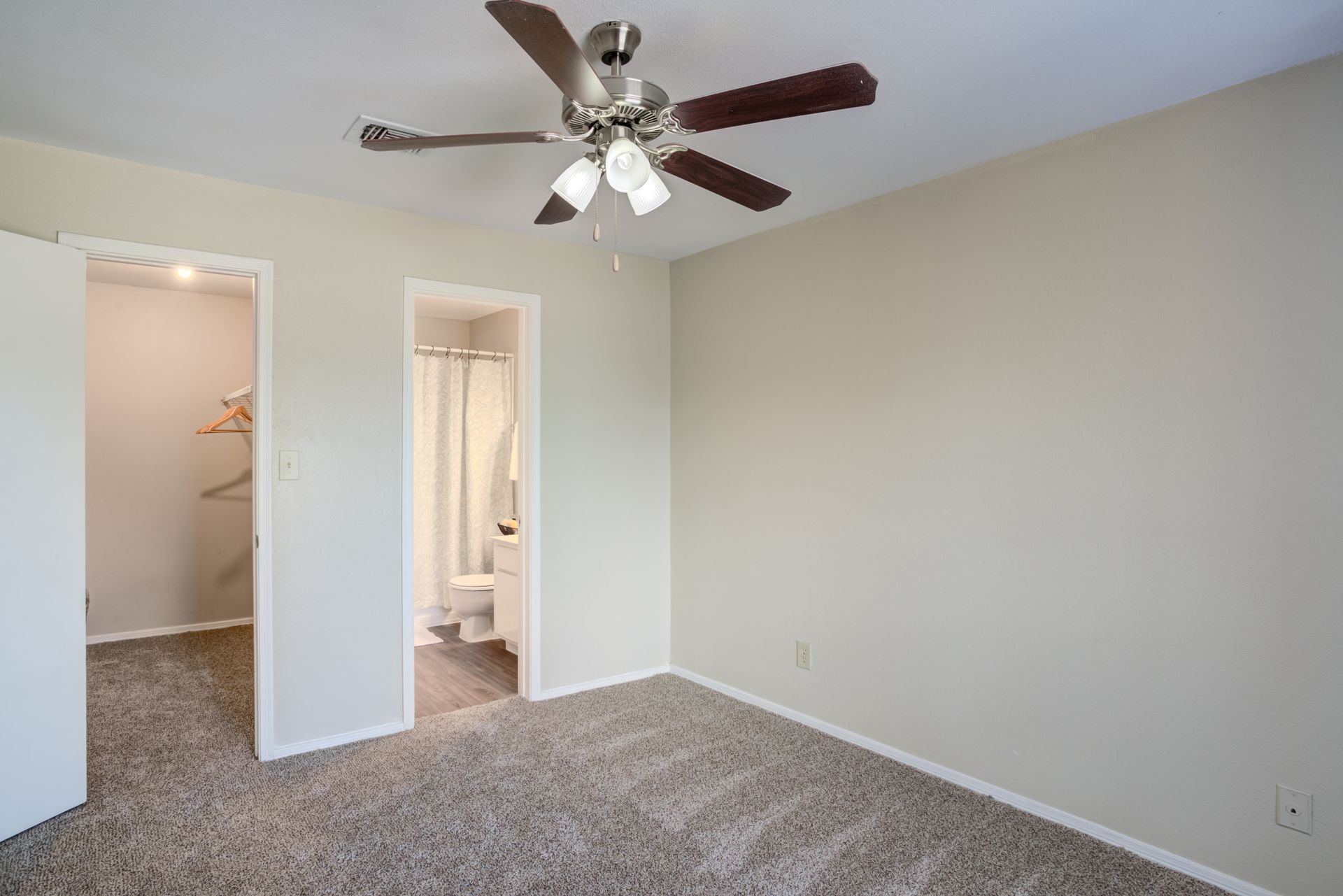 Bedroom with beige walls, carpet, ceiling fan, open closet, and bathroom doorway.