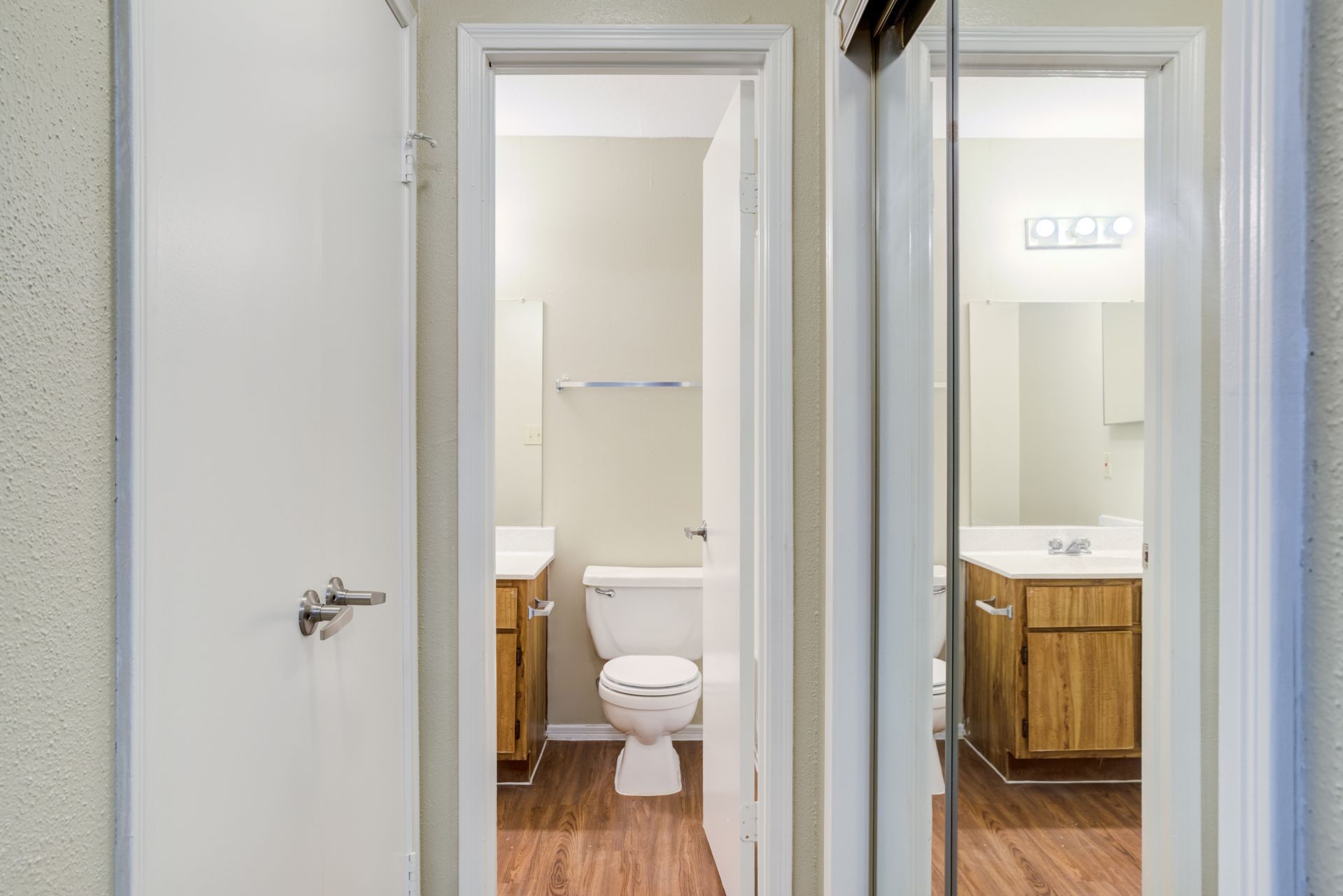 Open door to bathroom: toilet, sink, mirror, wooden cabinets, and floor.
