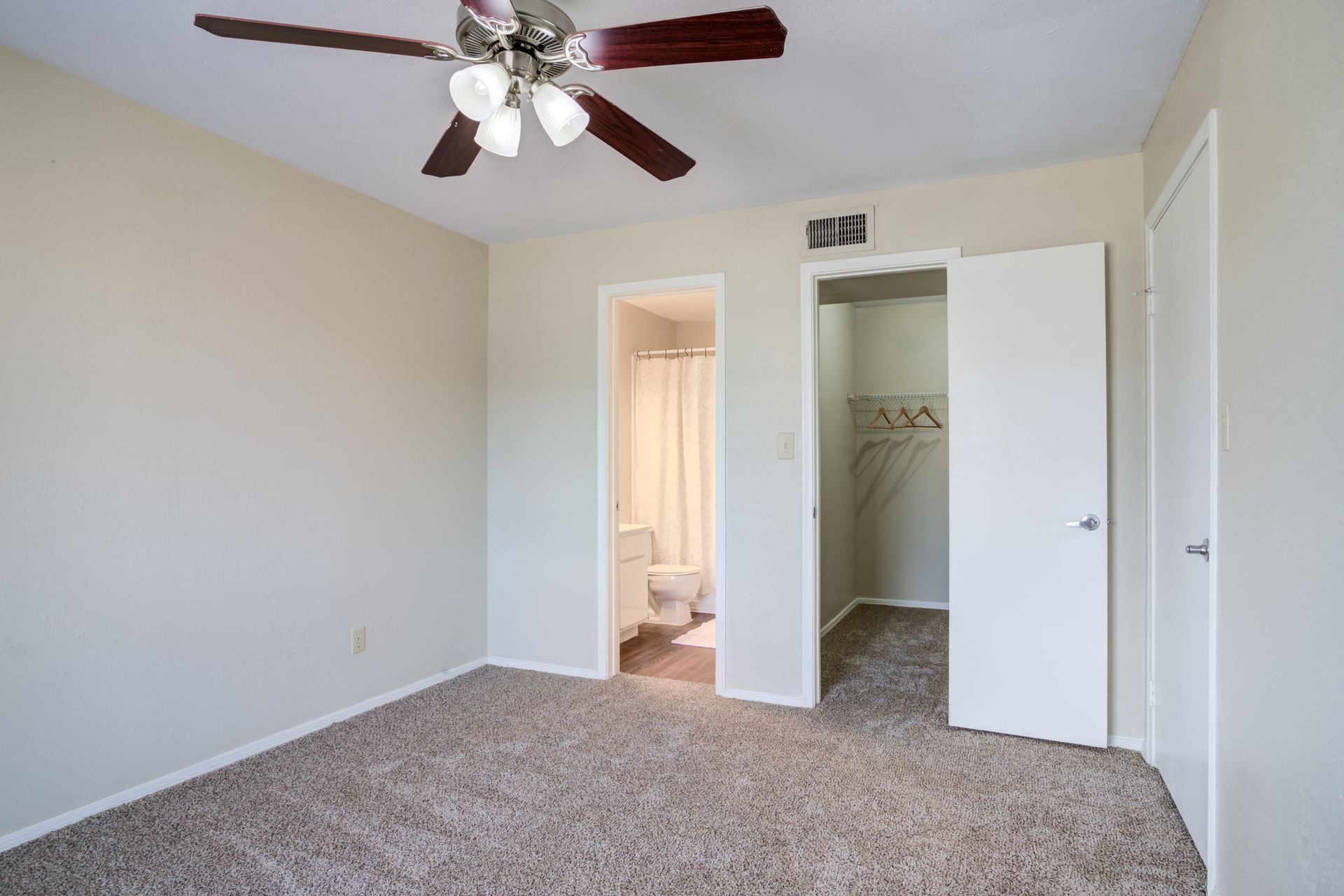 Bedroom with beige walls, brown carpet, ceiling fan, open closet, and doorway to bathroom.
