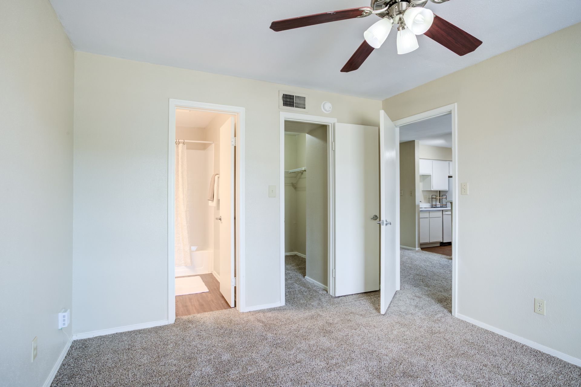 Bedroom with beige walls, carpet, and three white doors leading to bathroom, closet, and kitchen. Ceiling fan.