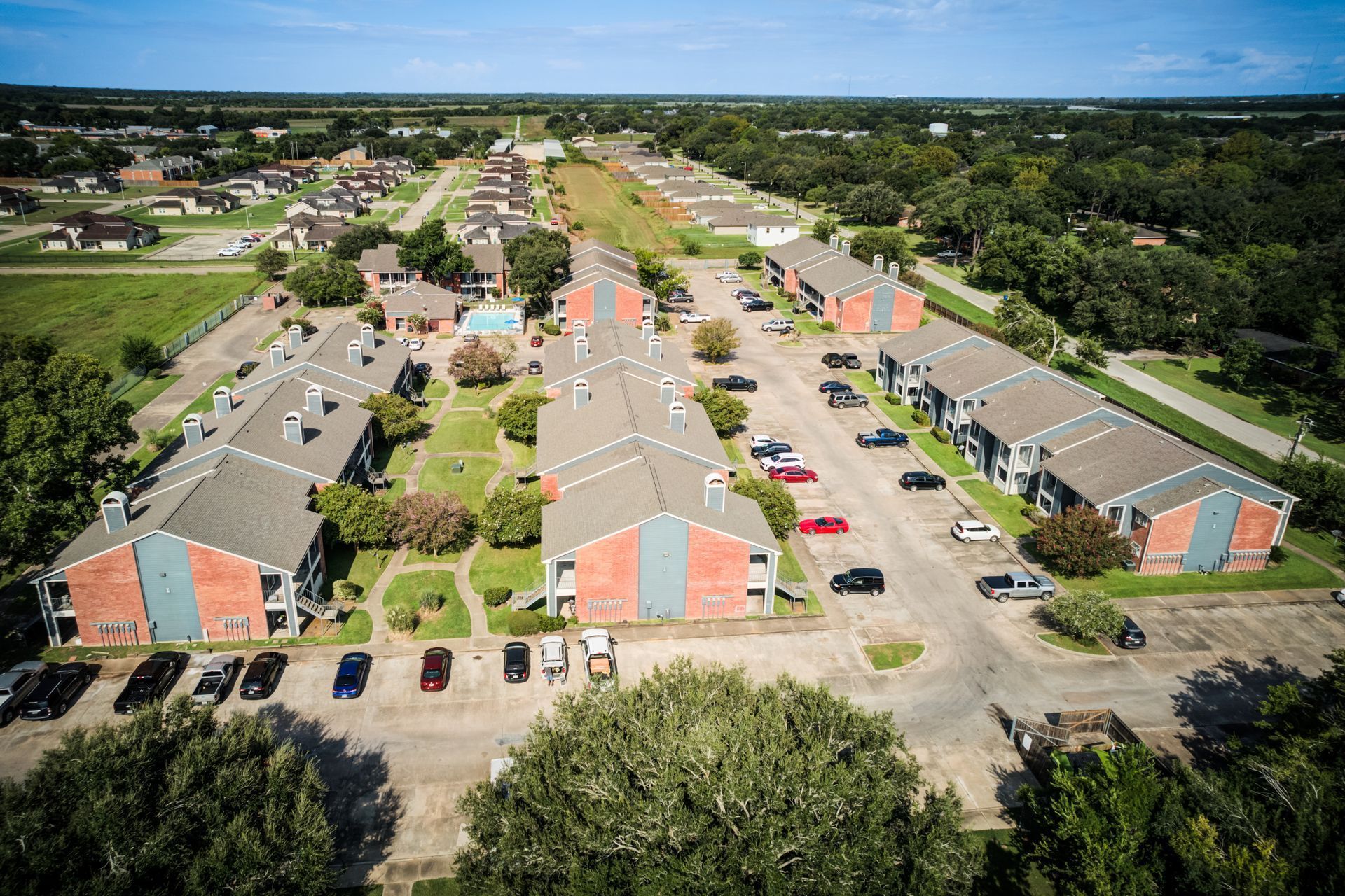 Aerial view of apartment complex with brick facades, parking, and a pool. Green lawns and trees surround the buildings.