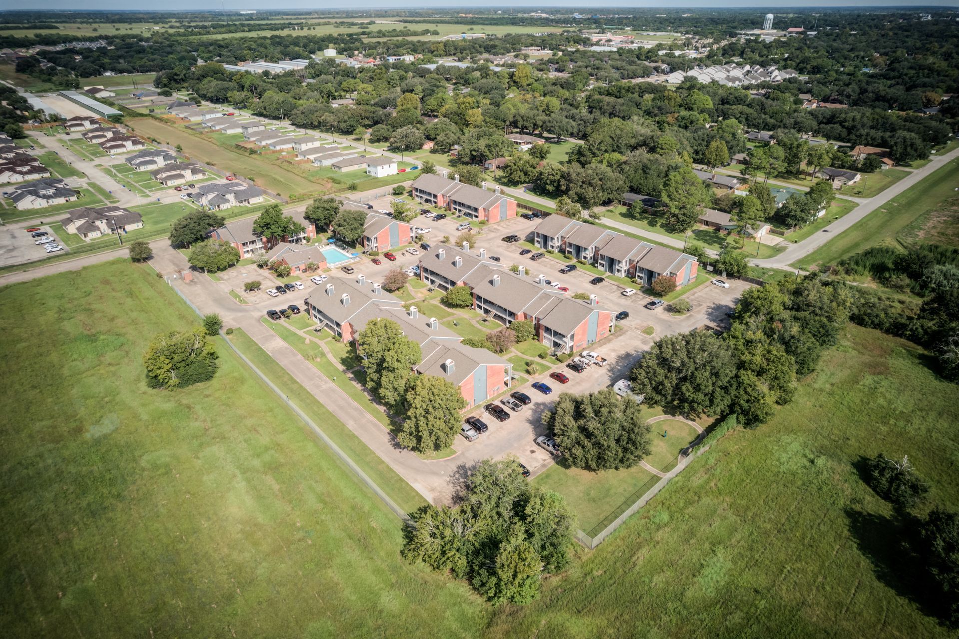 Aerial view of apartment complex with cars parked in front, surrounded by green fields and trees.
