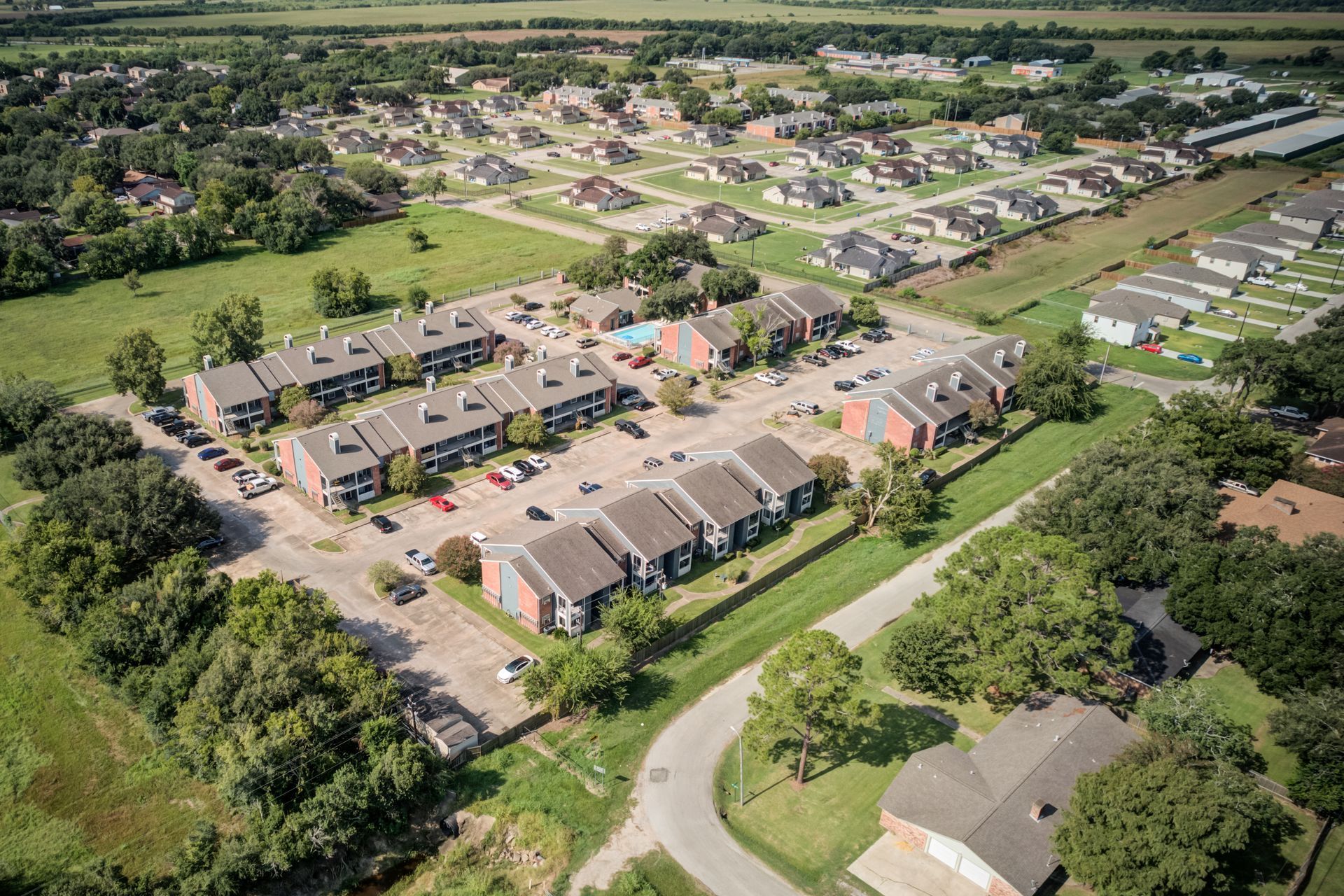 Aerial view of a suburban neighborhood with apartment buildings, a swimming pool, and houses surrounded by trees and green fields.