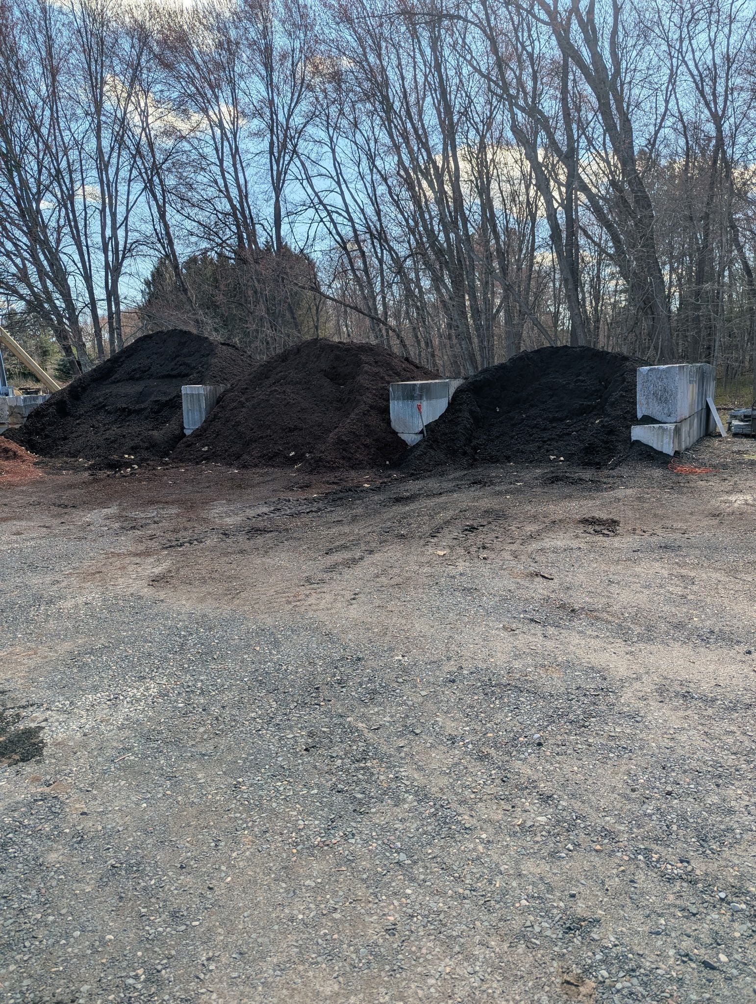 Three large mounds of dark mulch separated by concrete blocks on a gravel lot in front of a line of bare trees.