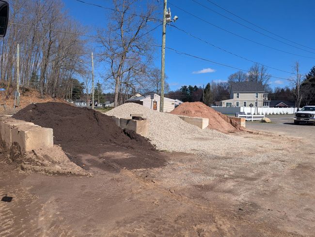 Three large mounds of landscaping materials, including dark mulch and gray gravel, held by concrete blocks in a yard.