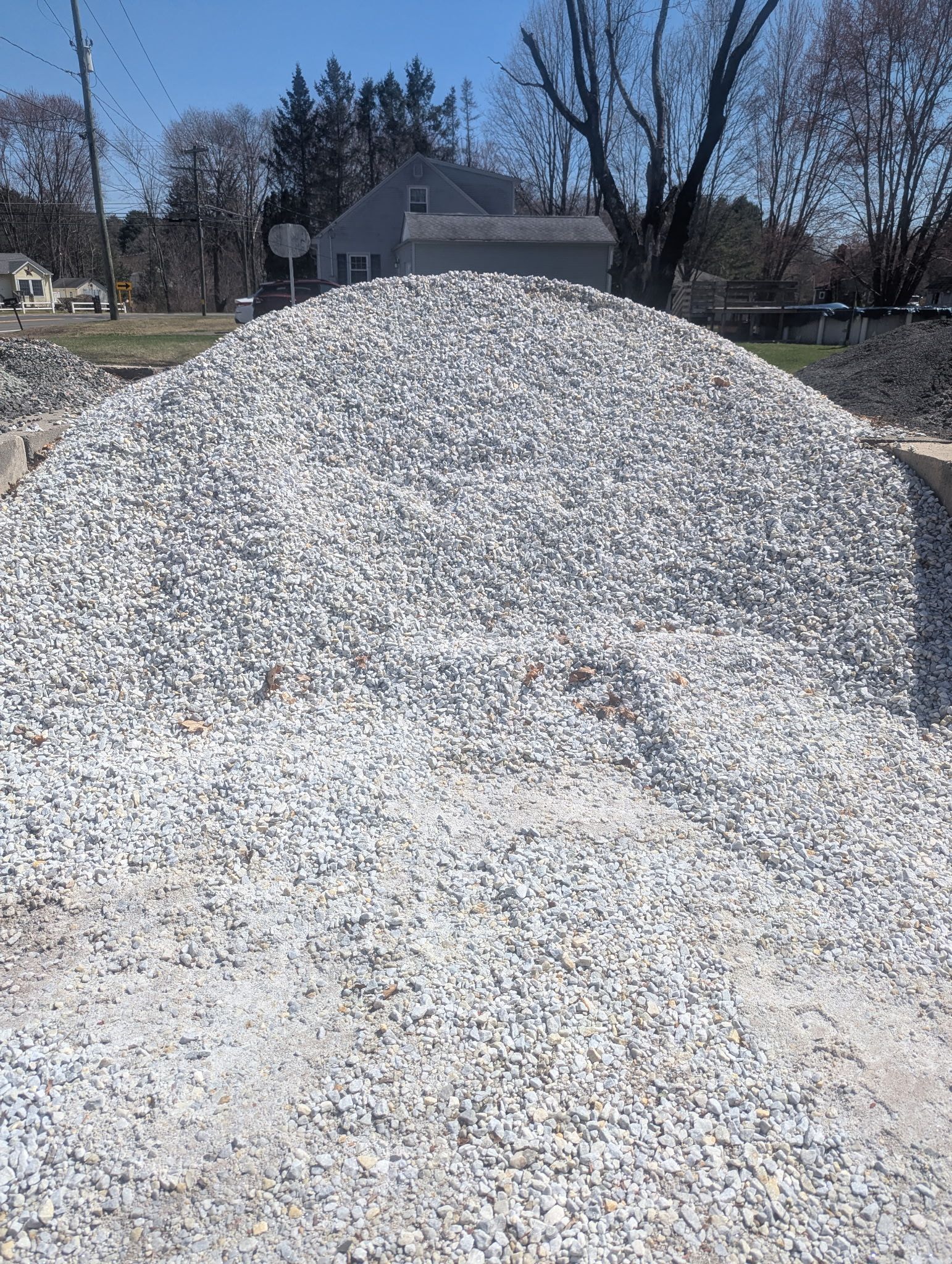 A large, conical pile of grey gravel outdoors under a bright blue sky, with a house visible in the background.