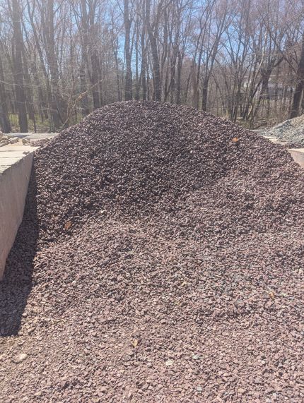 A large, conical pile of dark brown crushed landscaping stone outdoors against a background of bare trees.