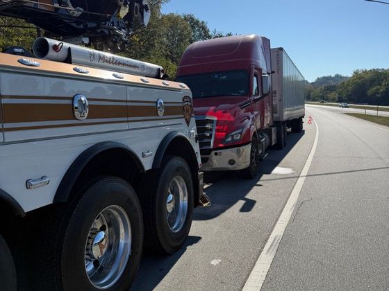 Tow truck towing a red semi-truck with a trailer on the side of a highway on a sunny day.