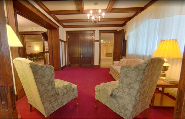 A sunlit room with dark wood trim, red carpeting, two patterned armchairs, a couch, and a matching table lamp.