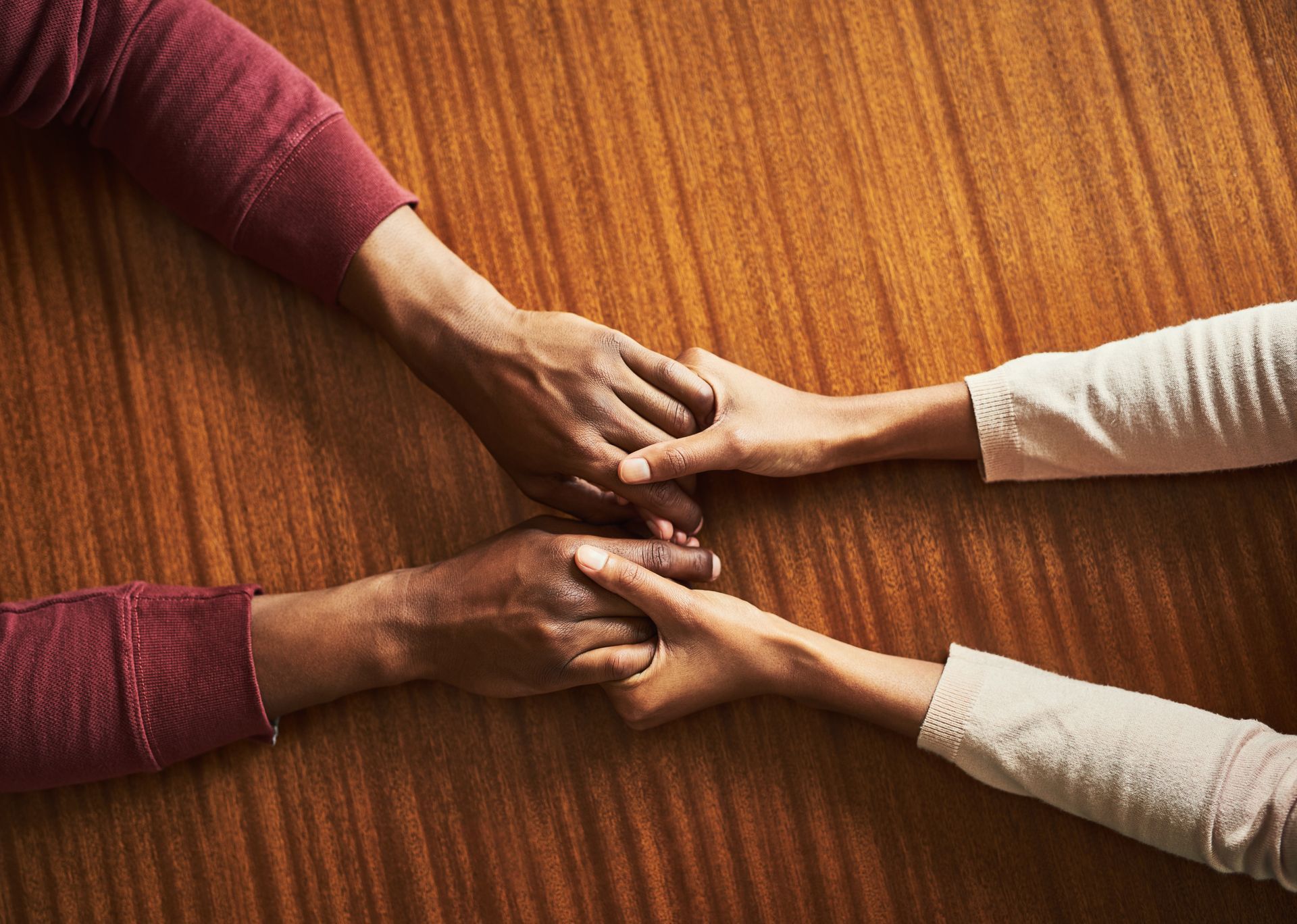 Two people hold hands on a wood-grained table, one person wearing a burgundy long-sleeved shirt and the other in beige.