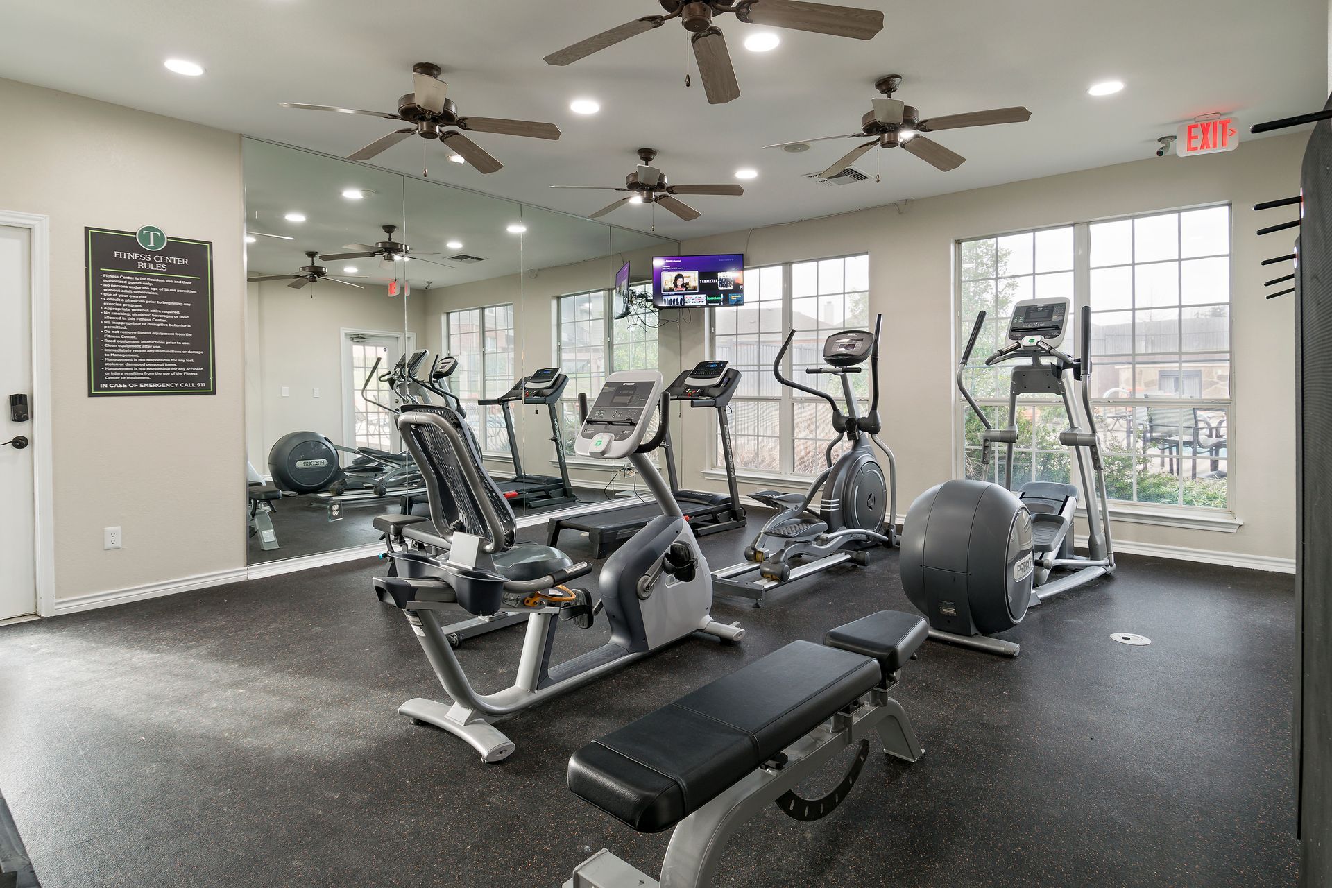 Gym with exercise equipment: treadmills, ellipticals, recumbent bike, weight bench, and ceiling fans.