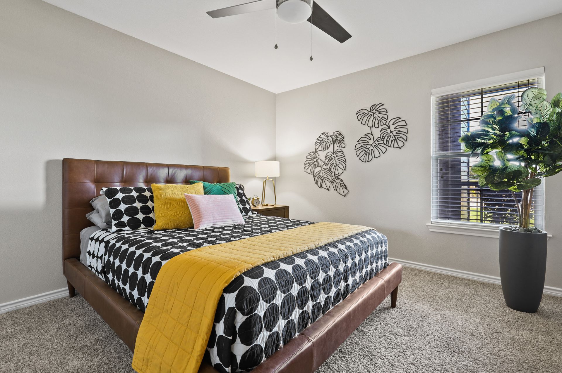 Bedroom with patterned bedding, leather headboard, small side table, and large houseplant near window.