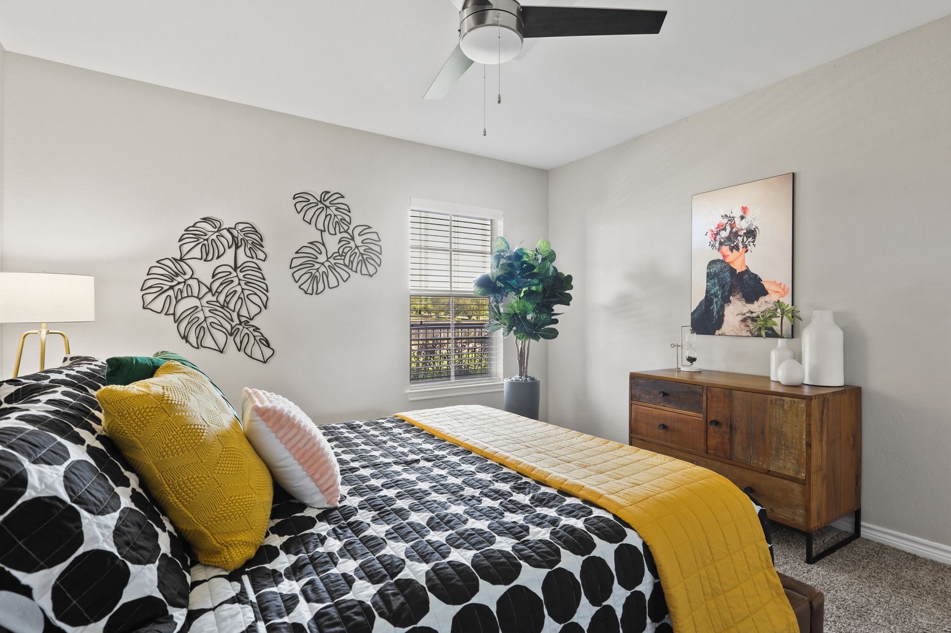 Bedroom with a bed featuring black and white patterned quilt, yellow throw and pillows, artwork, and a dresser.